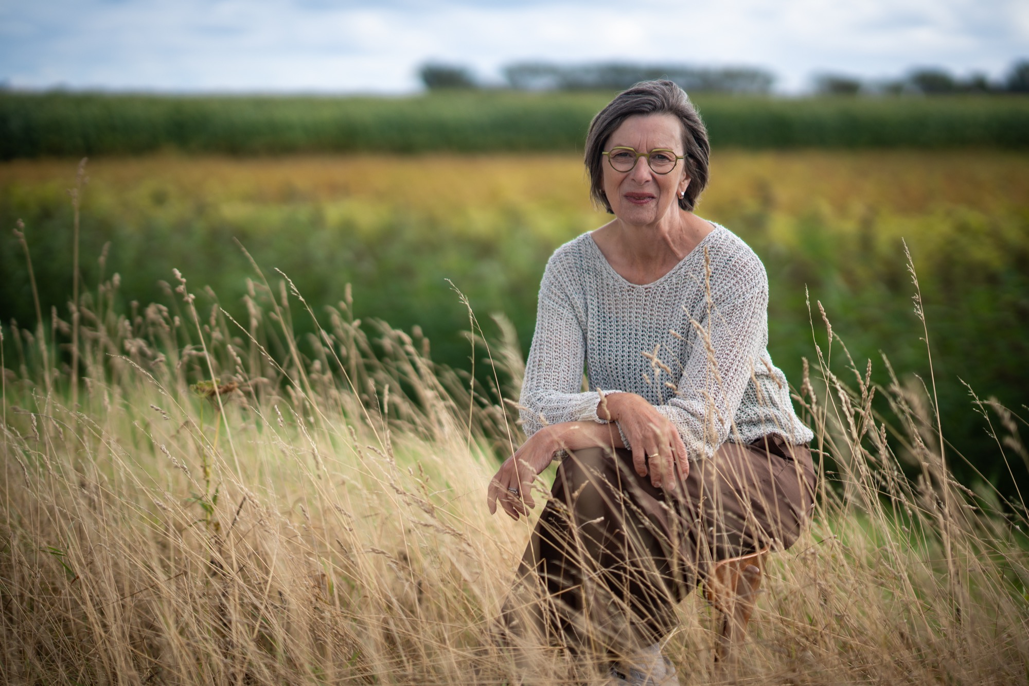 Rita van der Laag zit gehurkt in het hoge gras en kijkt in de camera